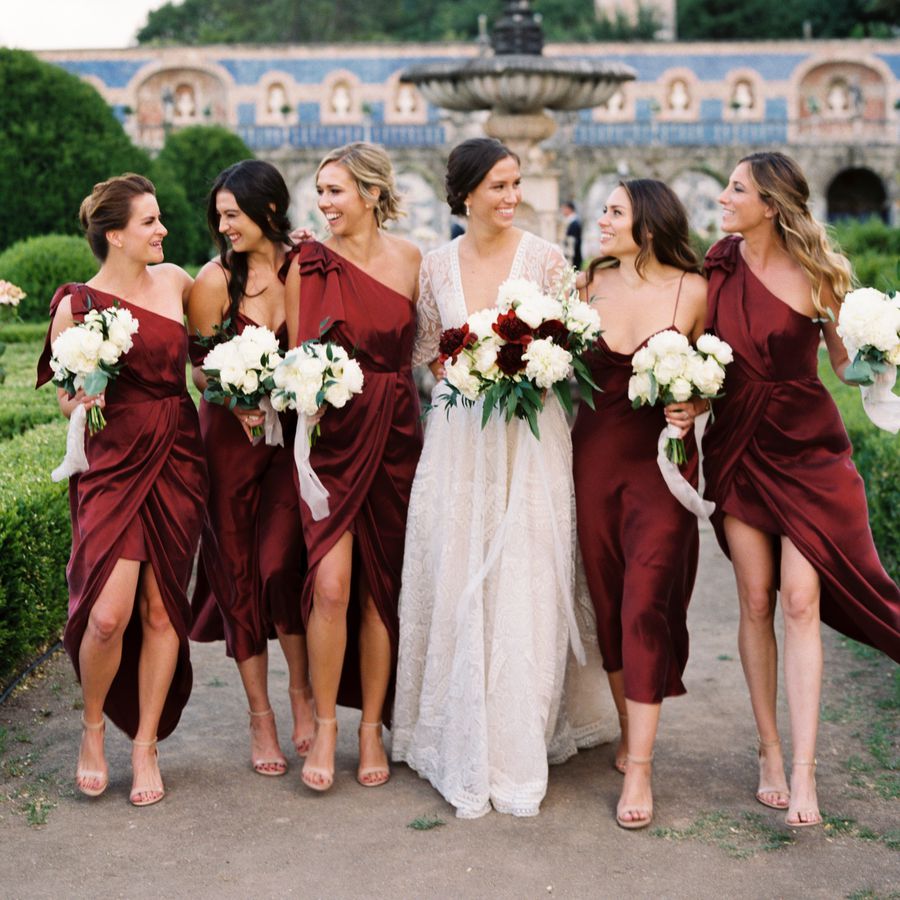 Bride surrounded by bridesmaids in burgundy dresses while walking in garden with nearby water fountain