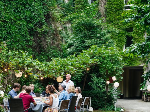 Family dinner outside of ivy-covered building in a courtyard with family members seated around table while father gives toast