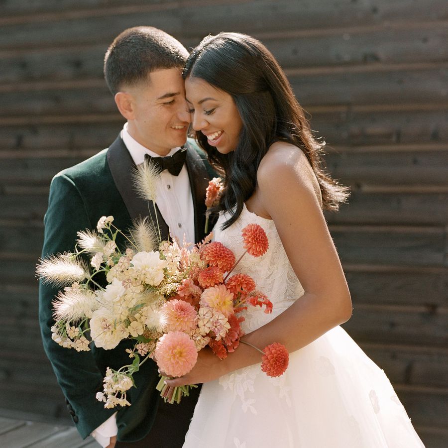 Bride holding a bouquet of terracotta dahlias, white hydrangea, and grasses while posing with the groom