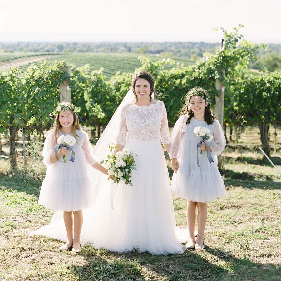 Person in a bridal gown holding the hands of two young people wearing matching formal blue gowns in a vast vineyard 