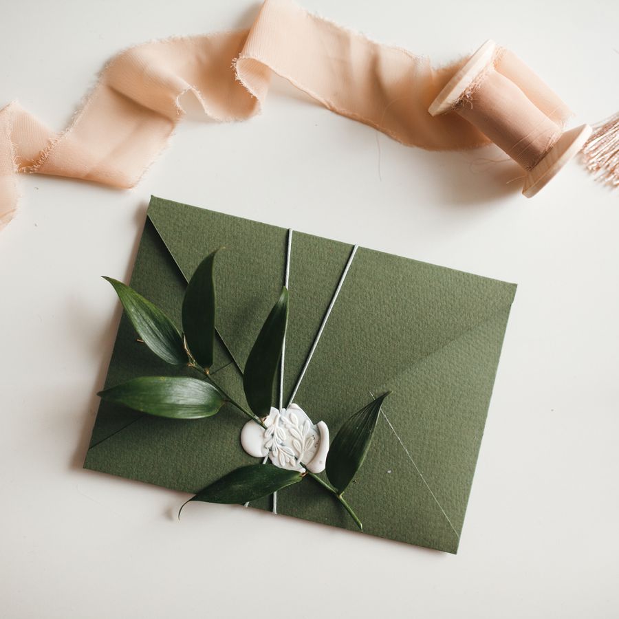 Green envelope wrapped with string and affixed with greenery on a white table with pink ribbon